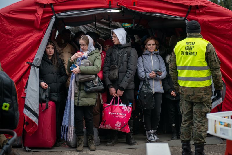 A group of Ukrainian refugees, bundled in warm clothing, stand under a red tent