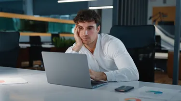 Man in a white shirt looking tired, resting his head on his hand while sitting at a desk with an open laptop in an office.