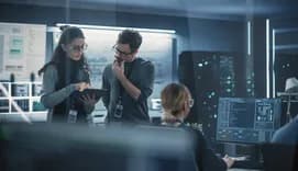Two people in glasses discuss artificial intelligence software on a tablet computer in a high-tech office, with computer monitors and a whiteboard in the background.