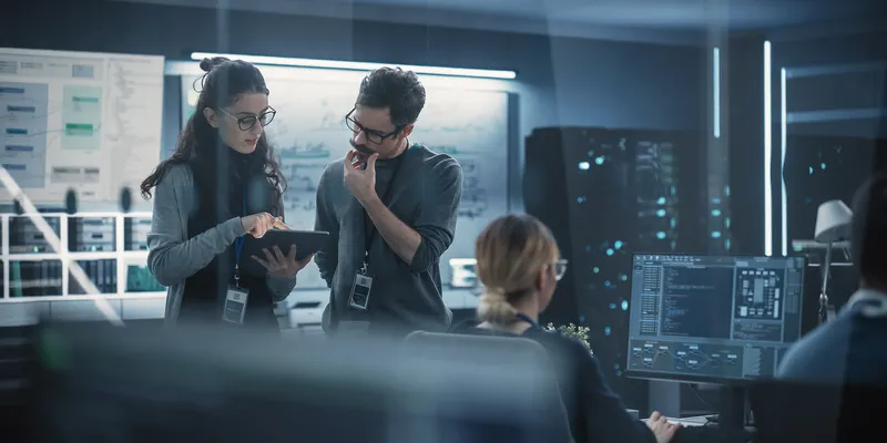 Two people in glasses discuss artificial intelligence software on a tablet computer in a high-tech office, with computer monitors and a whiteboard in the background.
