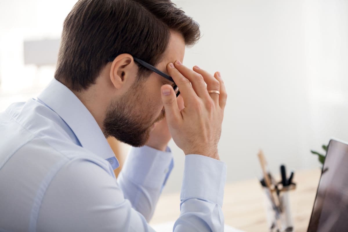 Man in a light blue shirt sitting at a desk, holding his head while looking at a laptop, appearing stressed or focused.