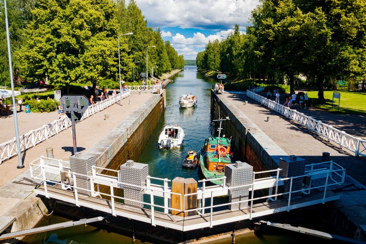 A canal lock with boats passing through, surrounded by green trees and pedestrians on the pathways under a partly cloudy sky.