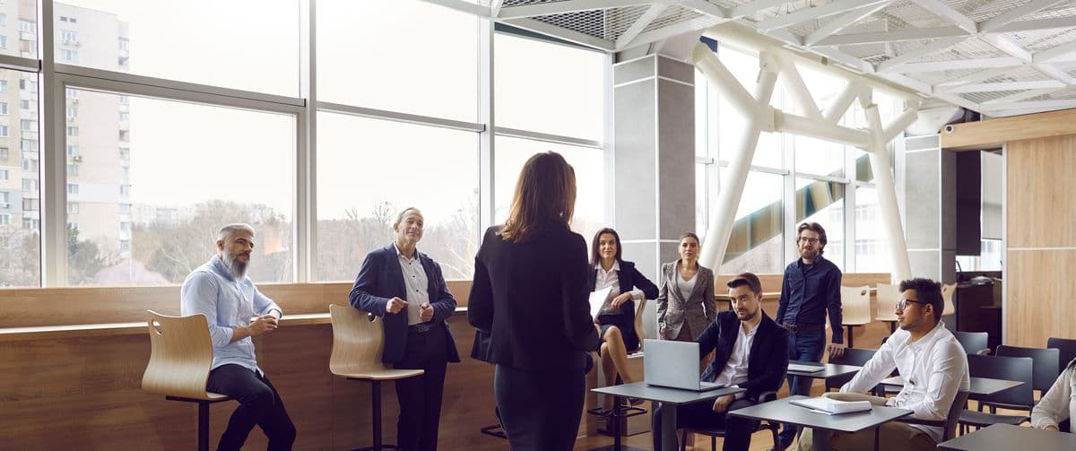 A woman stands addressing a group of seated colleagues in a modern office with large windows and natural light.