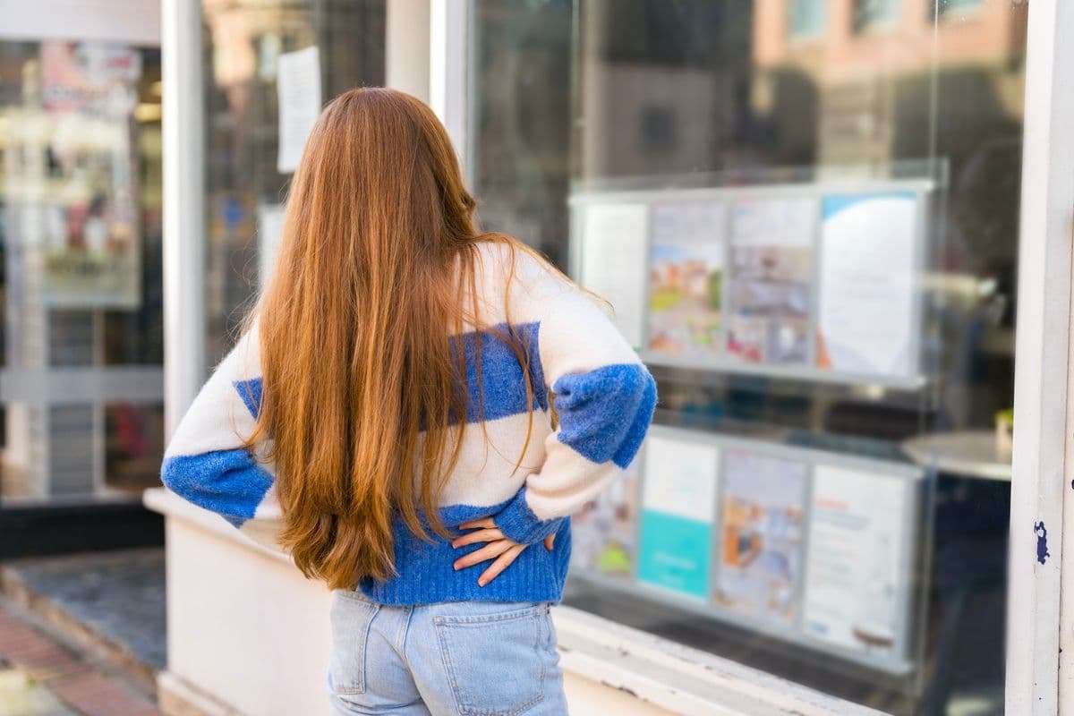 During daylight hours, a young woman examines real estate listings displayed in a shop window