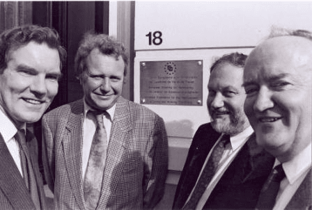 Four men in suits stand smiling in front of a building with a plaque and the number 18. The image is in black and white.