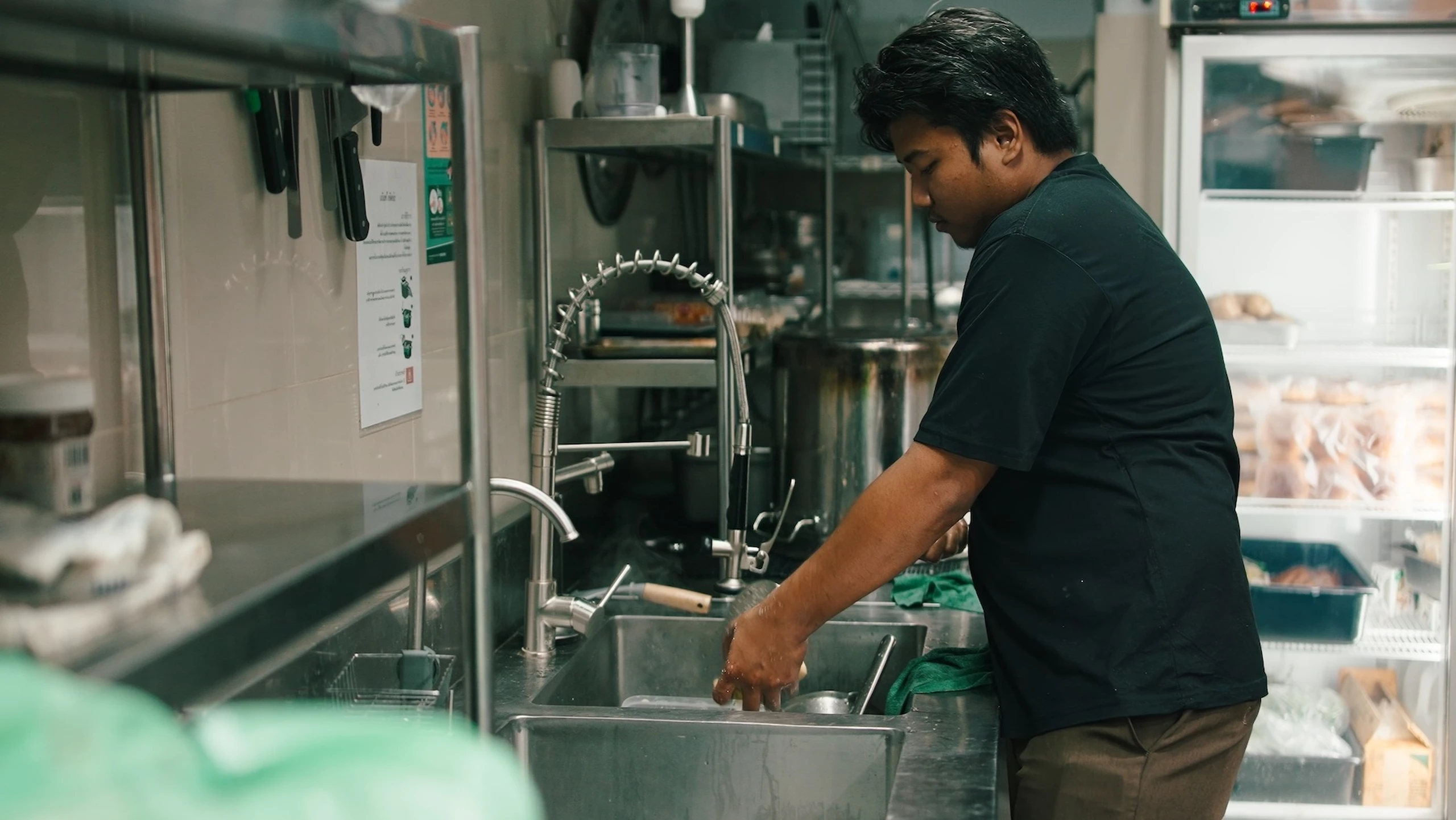 A person washing dishes in a commercial kitchen, standing near a double sink with shelves and equipment in the background.