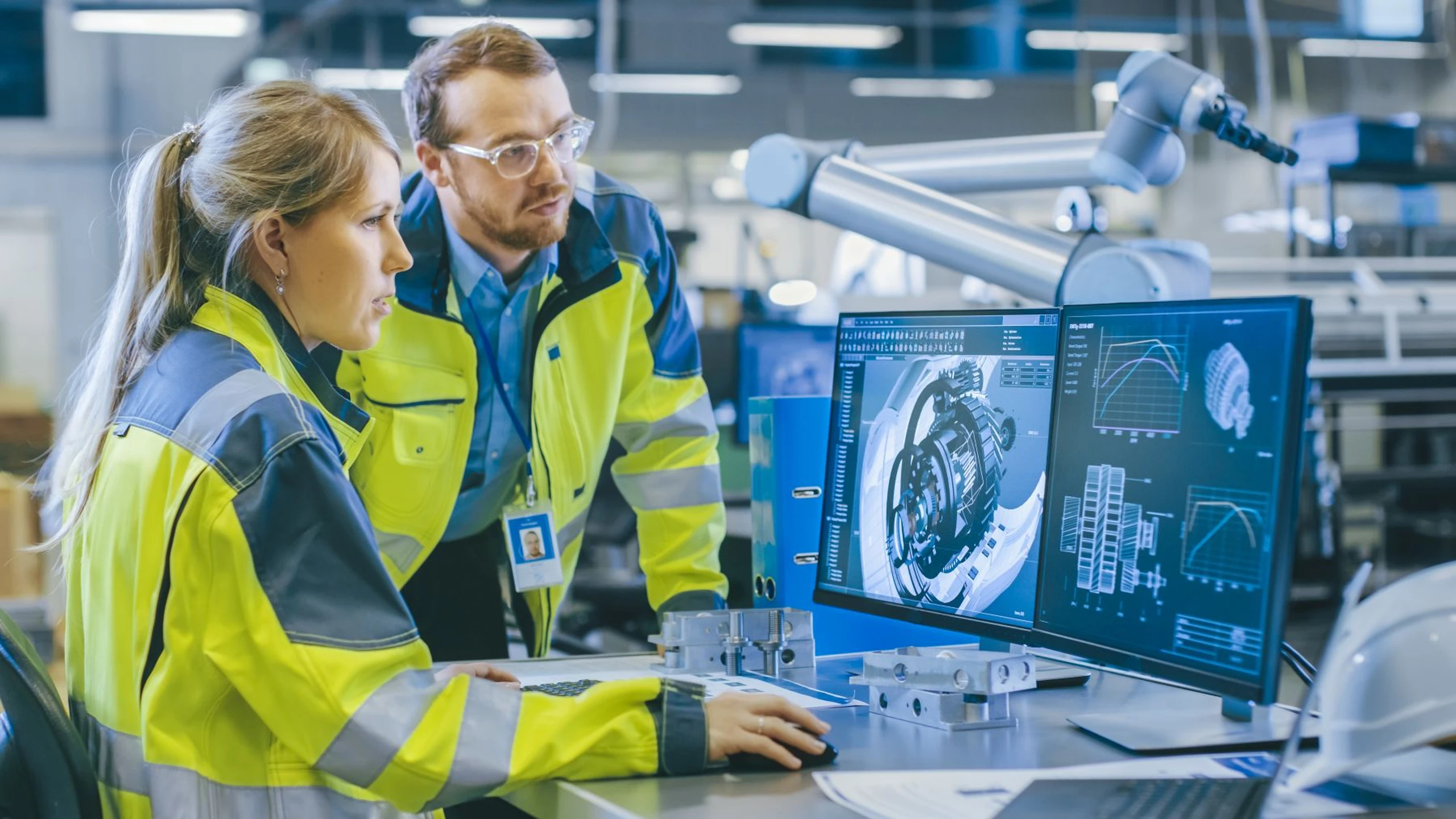 Two engineers in safety jackets analyze 3D mechanical designs on computer screens in a modern factory setting.