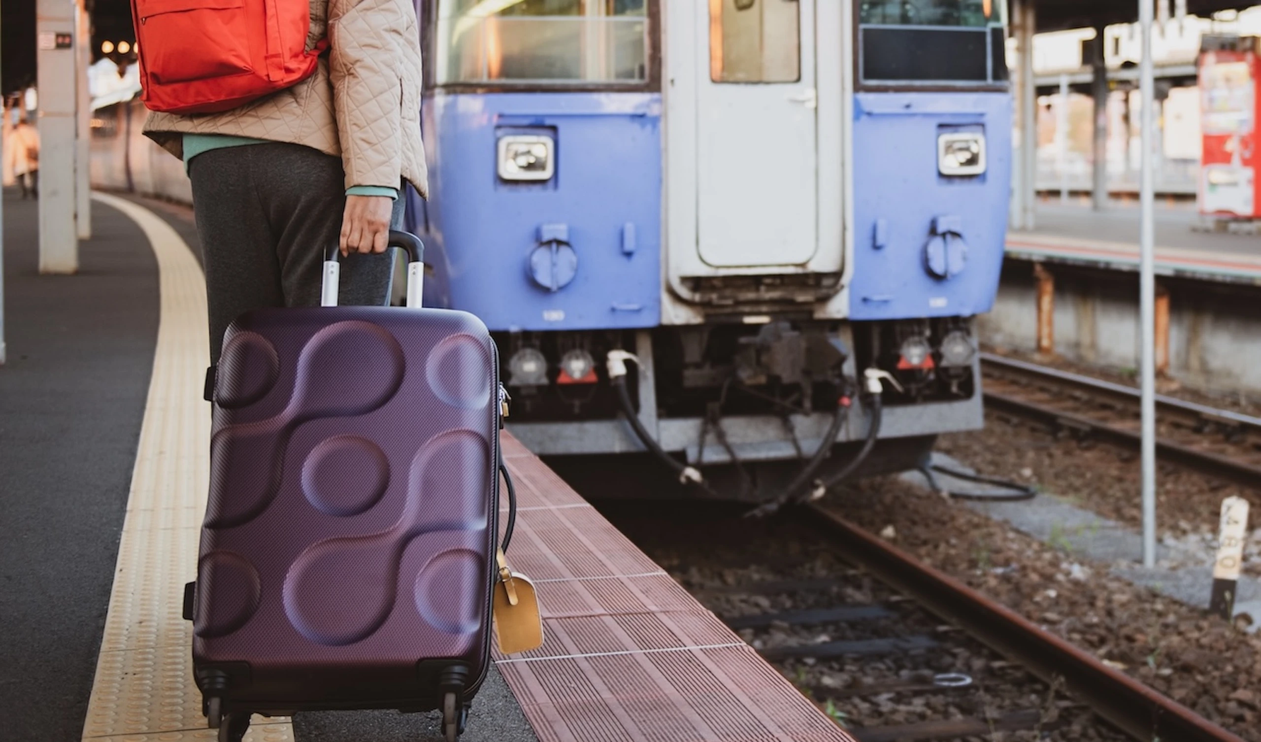 Person with a red backpack and purple suitcase waits at a train station platform beside a blue train.