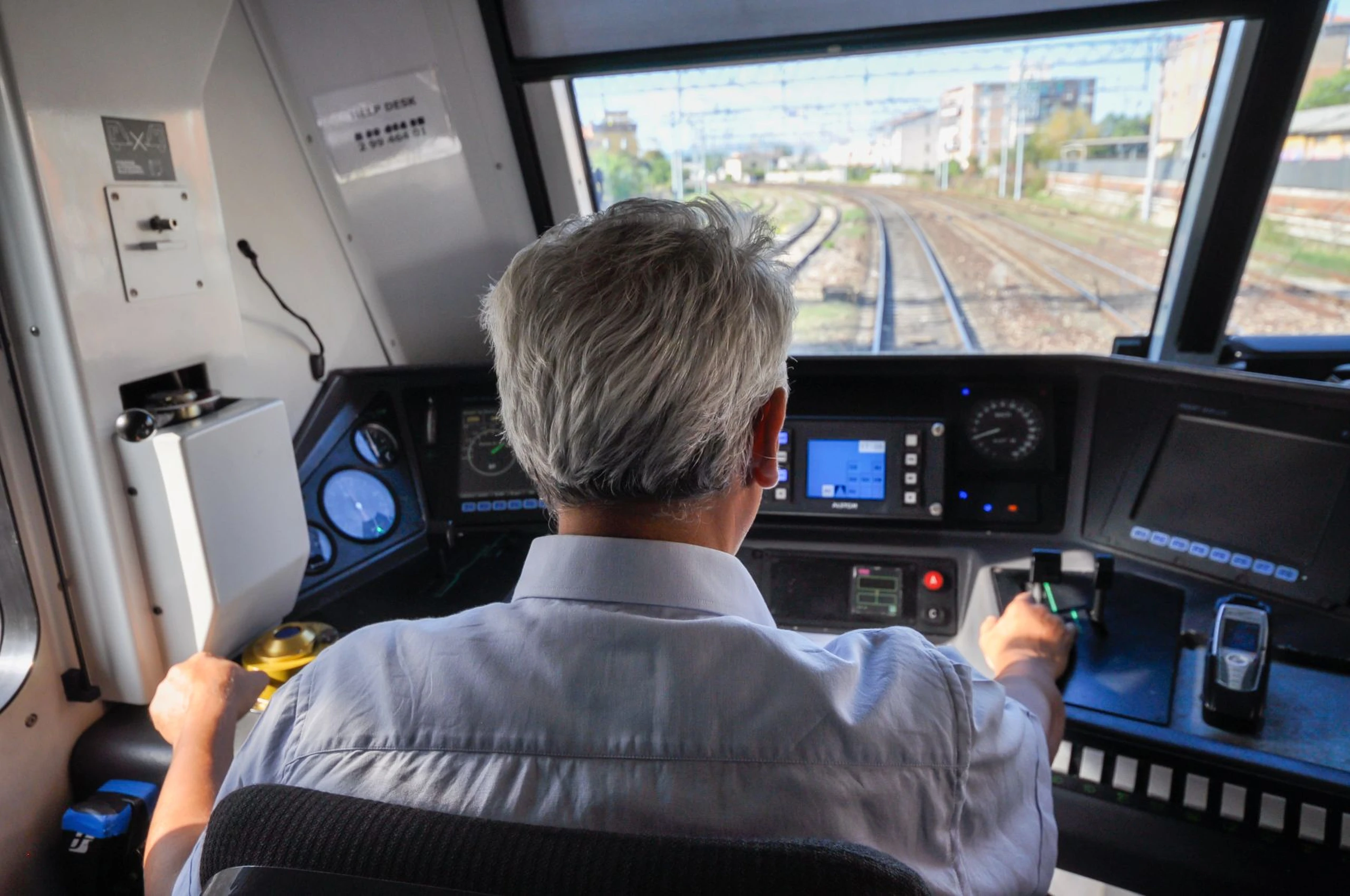 Train engineer operating control panel in a locomotive cab, looking out front window at railway tracks.