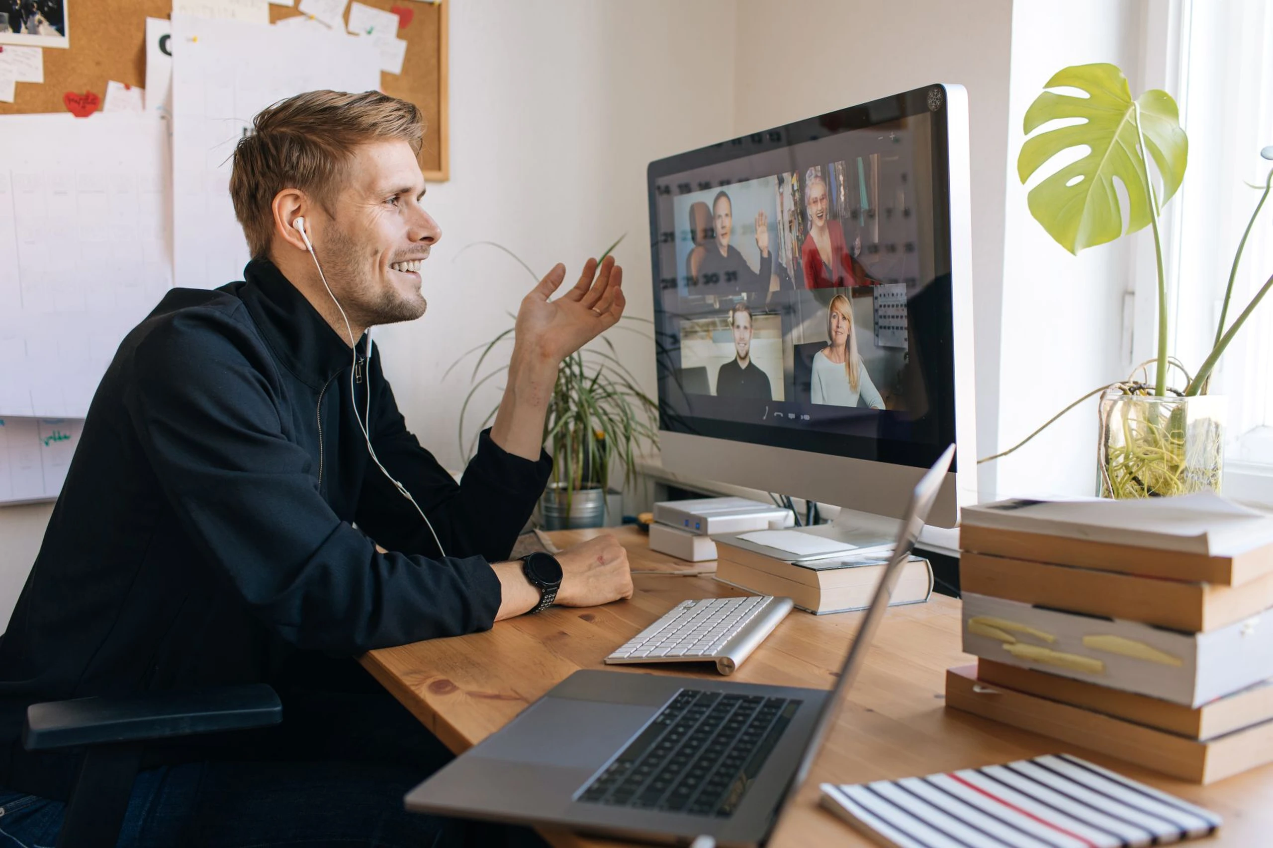 A man sitting at a desk, engaged in a video call on a computer, with books, a plant, and a laptop around him.