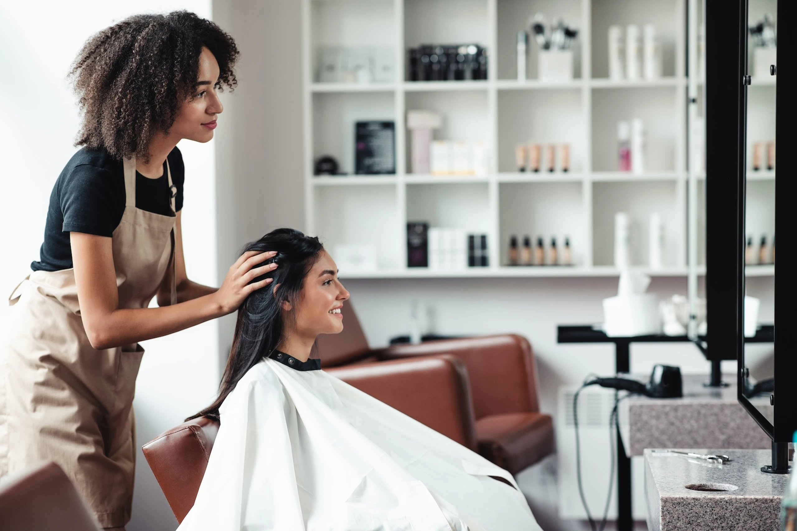 A hairstylist in an apron is working on a smiling client's hair in a salon with shelves of products in the background.