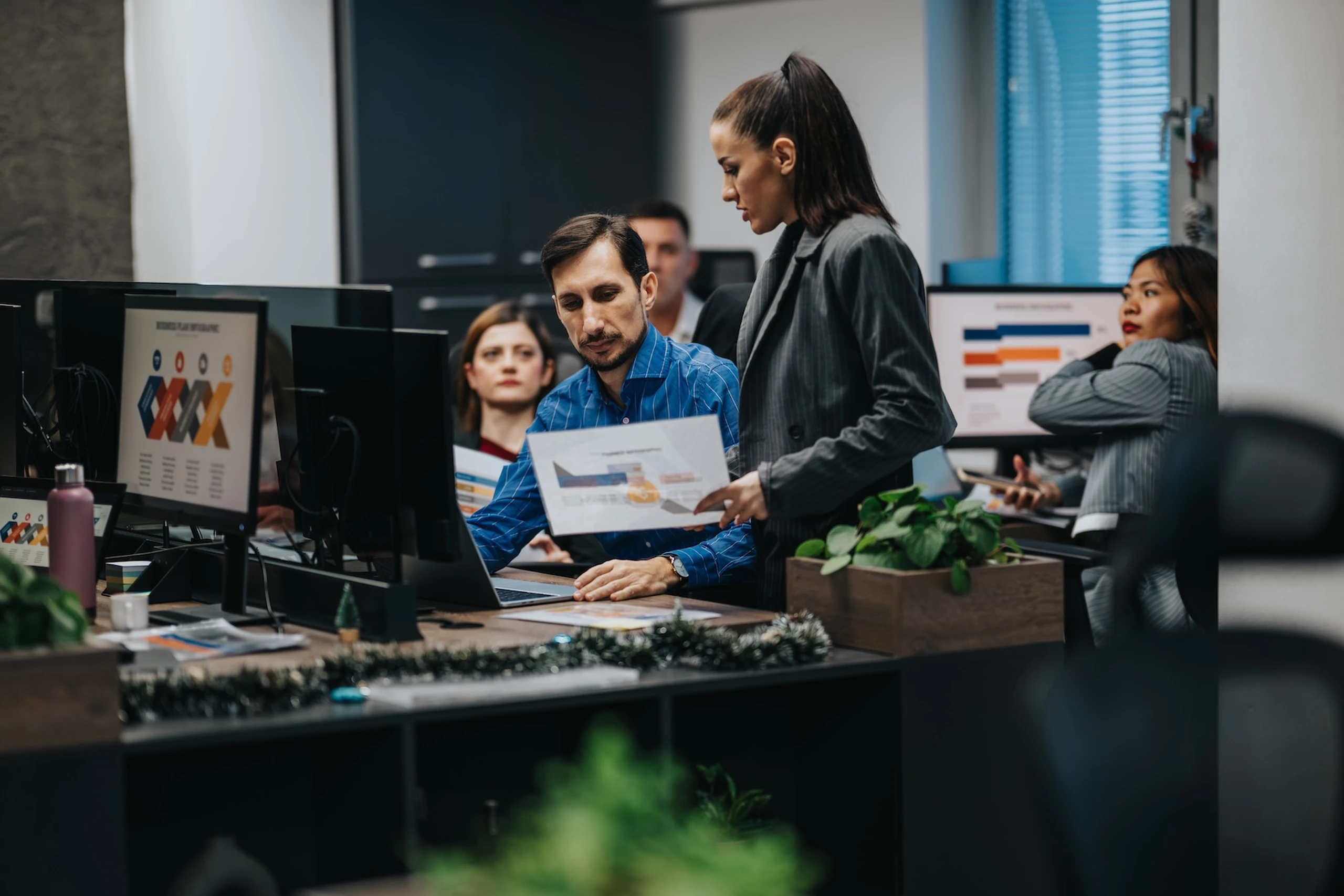 People collaborating in an office with computers and charts. A woman stands and shows a document to a seated man.