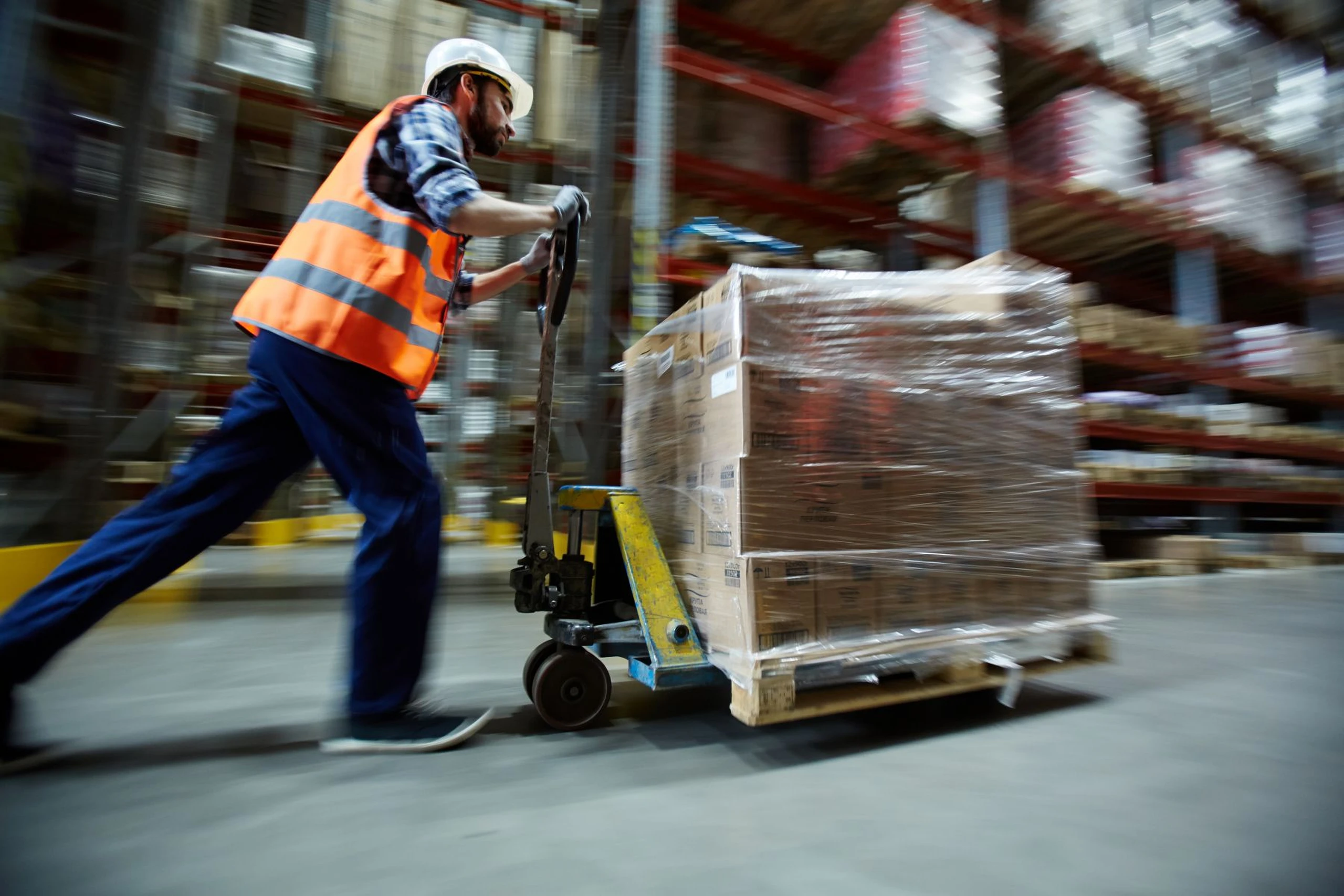 Worker in a high-visibility vest and hard hat moving a pallet with a hand truck in a warehouse.