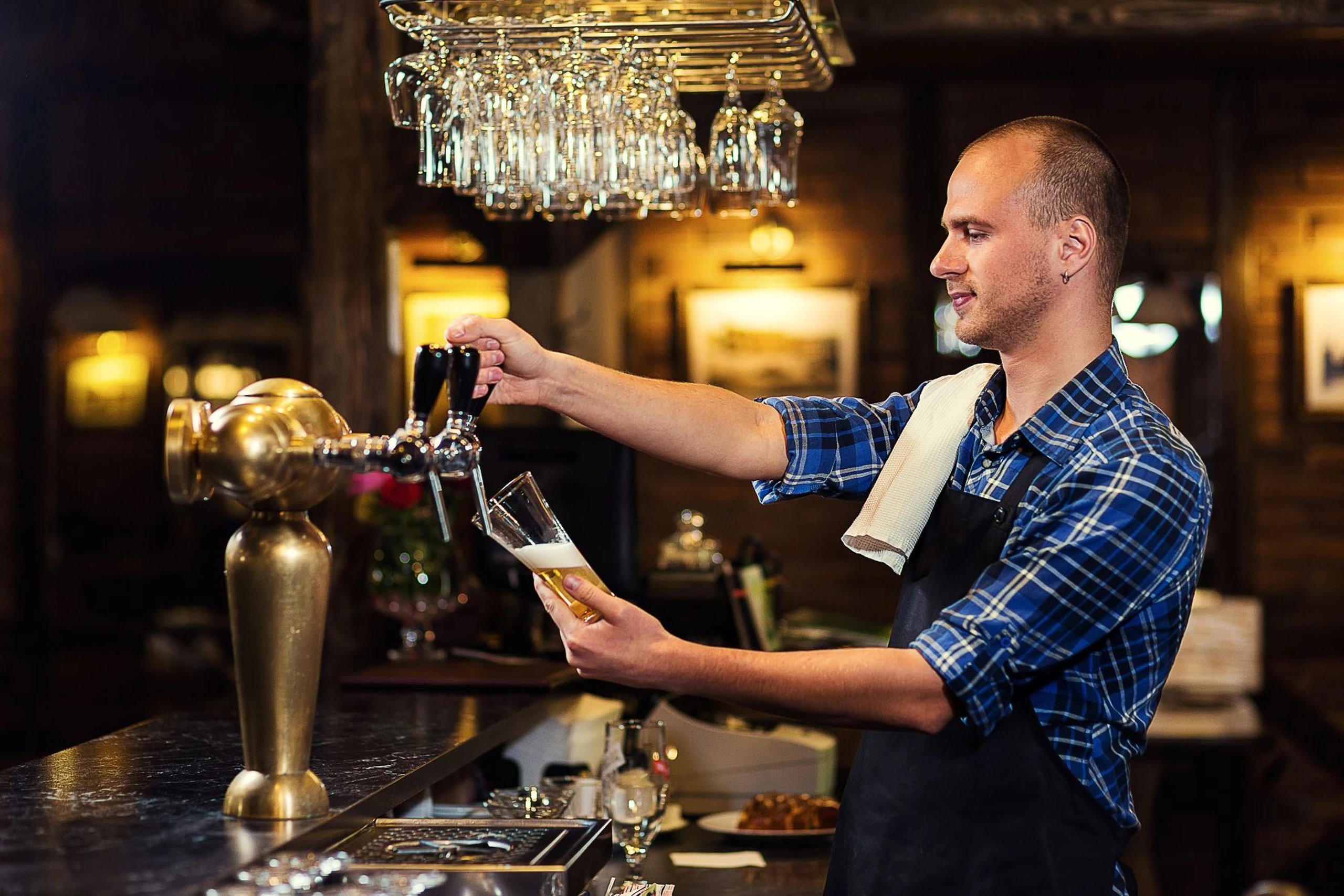 Bartender in a blue plaid shirt pours a beer from a tap at a dimly lit bar, surrounded by hanging glasses and wooden decor.