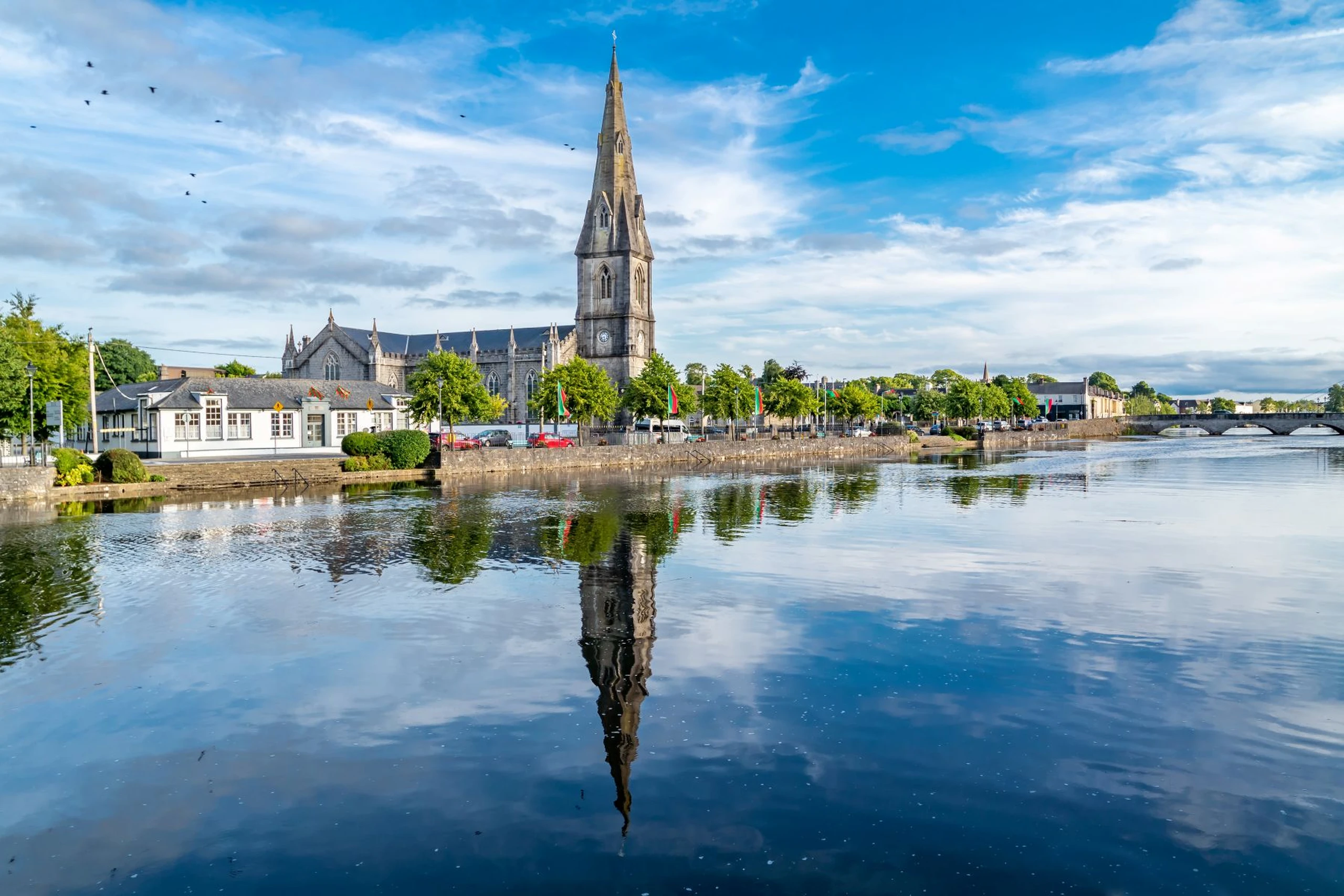 Tall church spire and buildings reflected in a calm river under a blue sky with scattered clouds and flying birds.