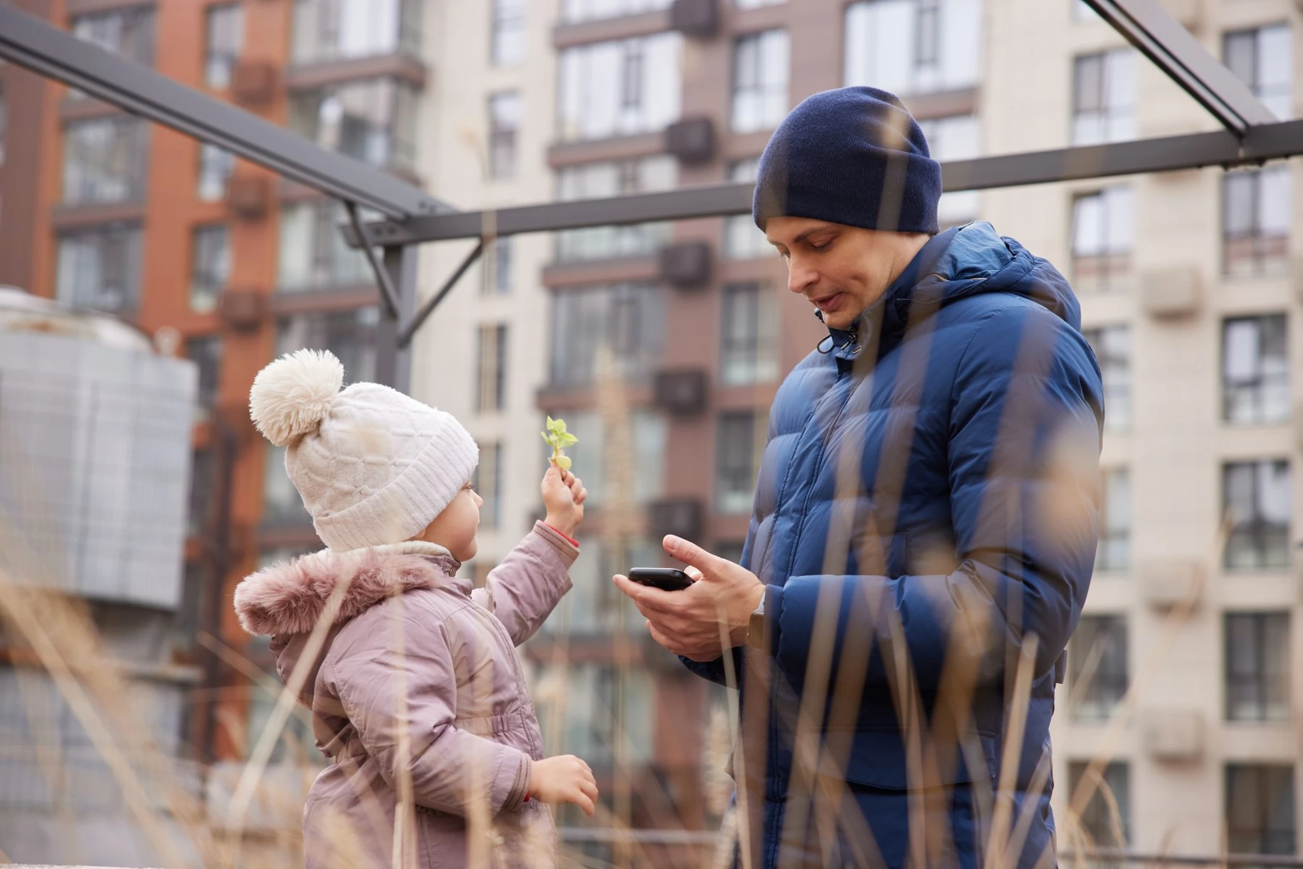 Child in a white hat offers a leaf to an adult in a blue jacket, standing with a phone in an urban setting.