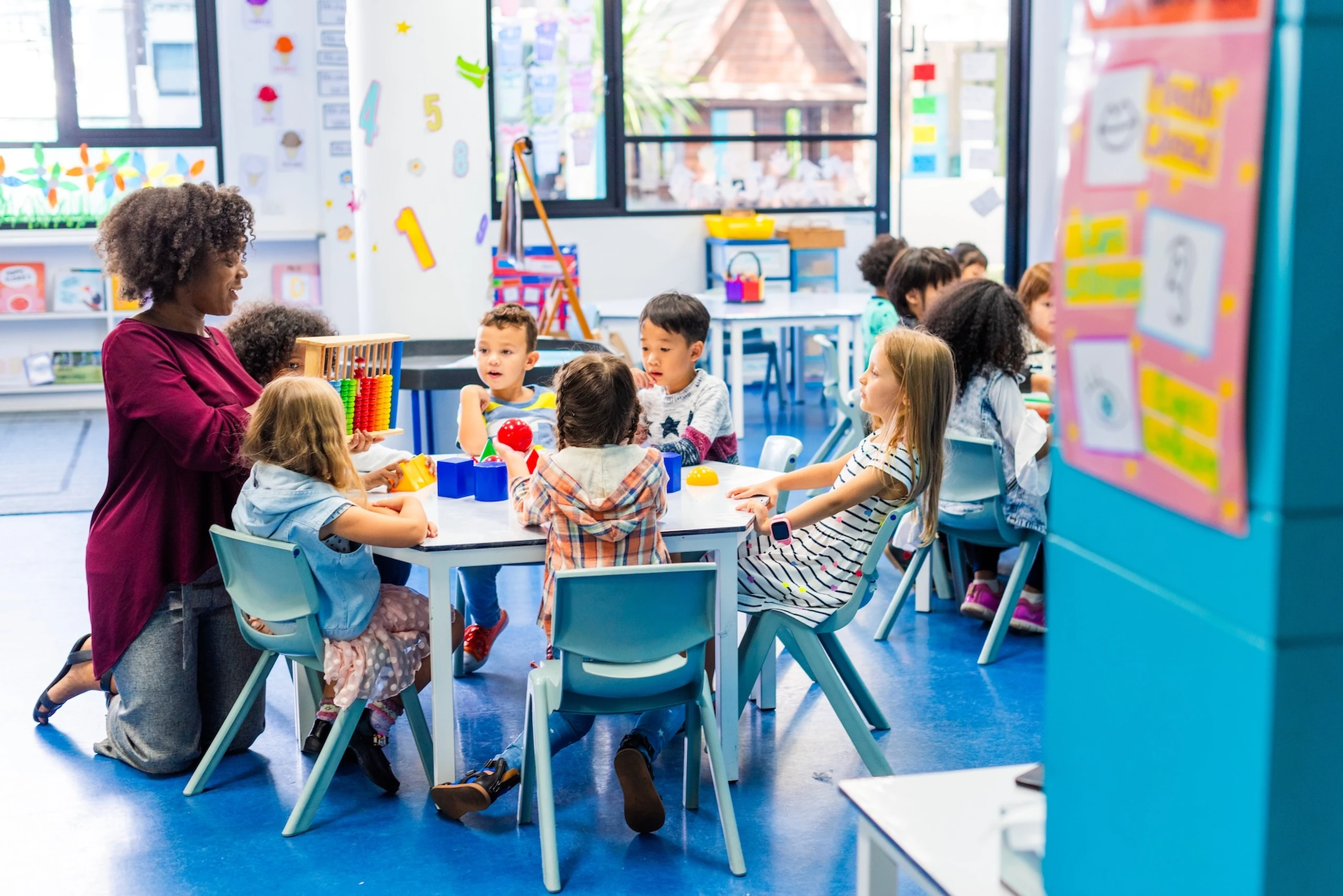 A teacher engages young children seated around a table with educational toys in a colorful classroom.