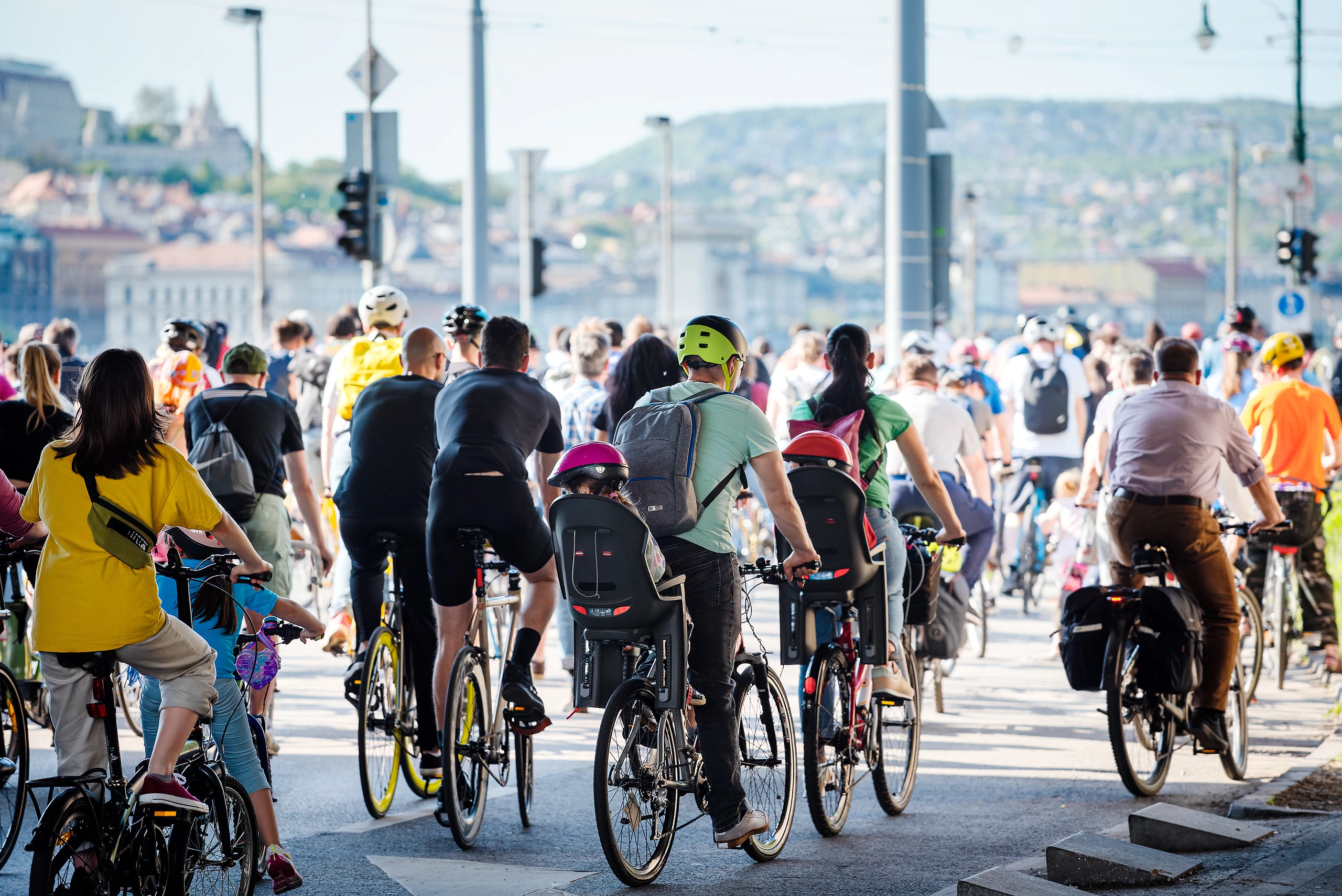 A large group of cyclists riding together in a city, some carrying children in rear seats; a scenic backdrop of hills and buildings is visible.