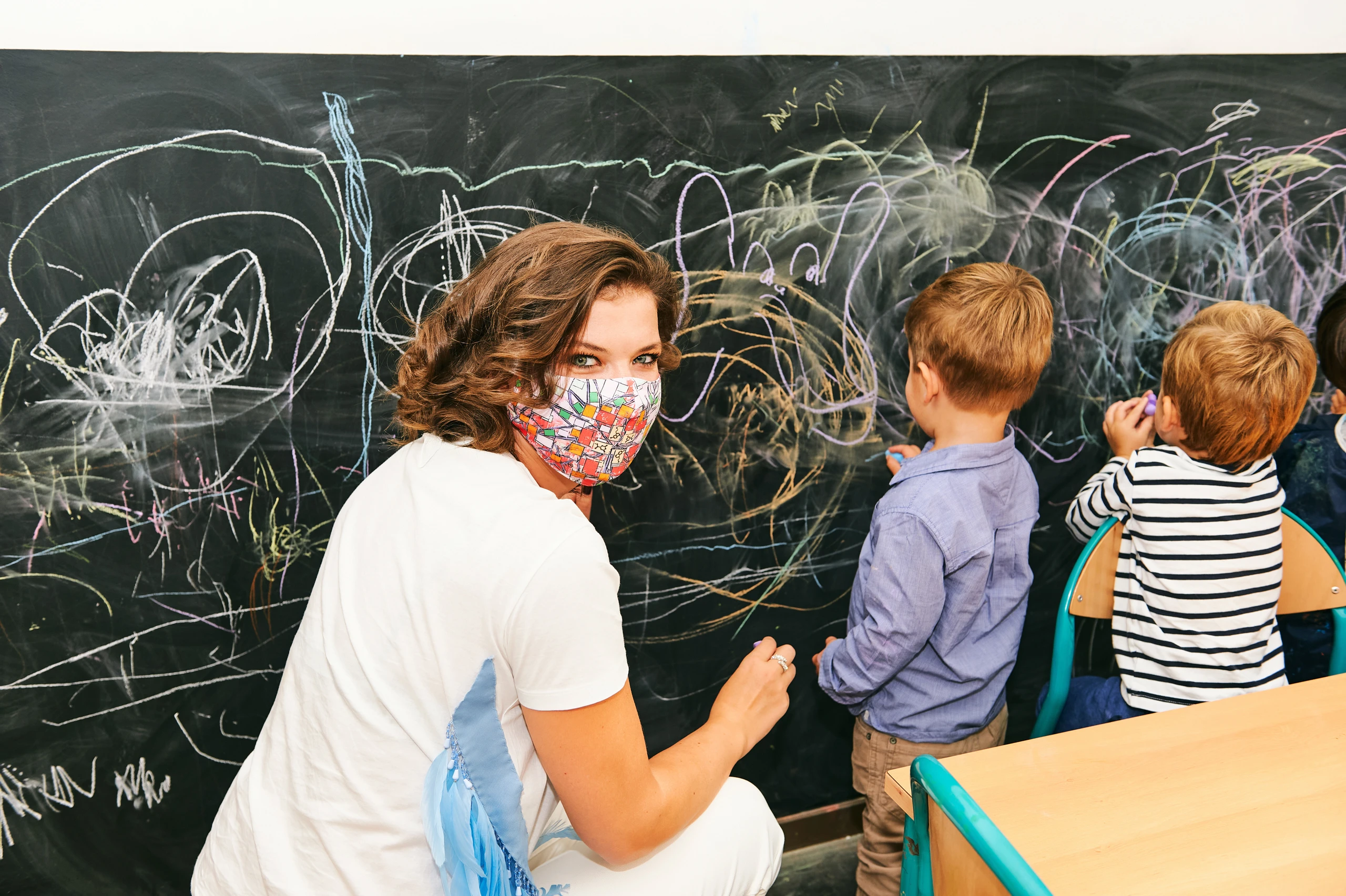 Art classroom in kindergarten, young teacher drawing with children on chalk wall, wearing facemask  by annanahabed / Adobe Stock