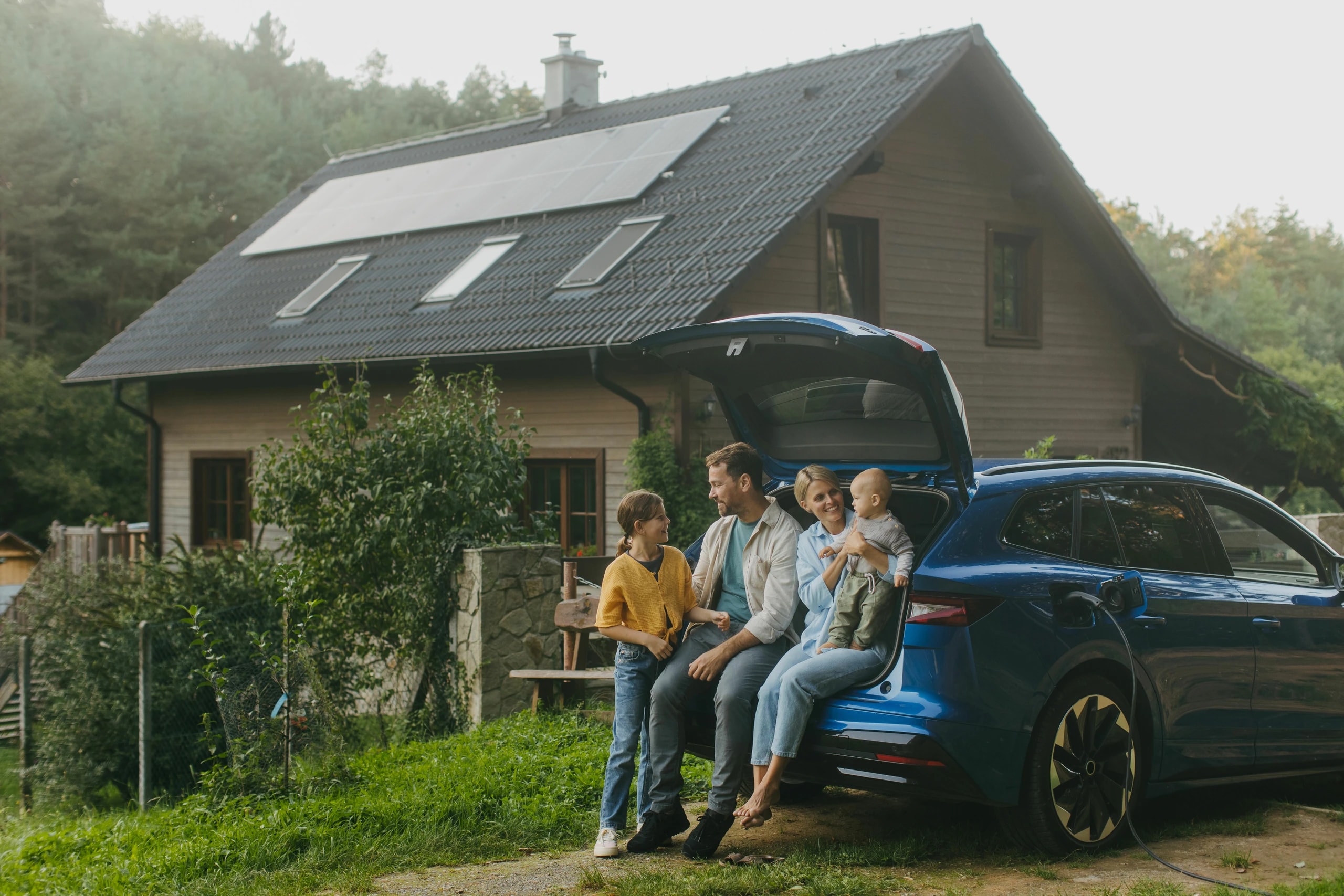 Family sitting on an electric car's trunk, in front of a house with solar panels, surrounded by trees.