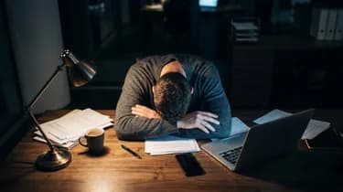 A person sits at a desk with their head resting on folded arms, surrounded by paperwork, a coffee cup, and a laptop, under a desk lamp.