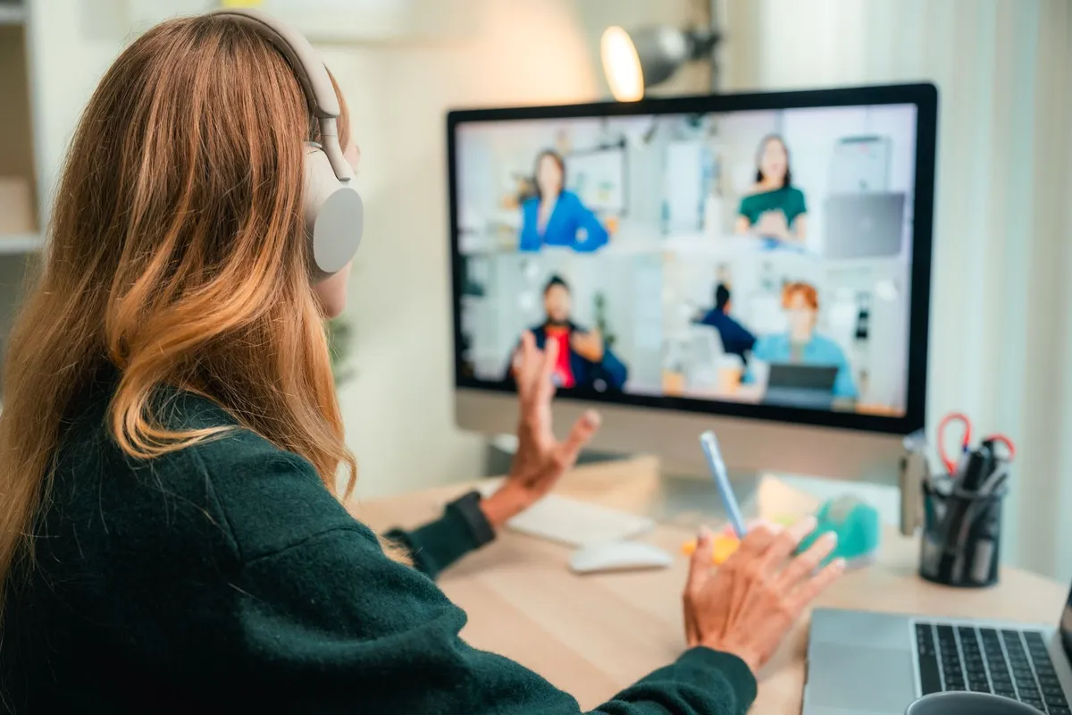A woman with headphones participates in a virtual meeting on a computer screen, sitting at a desk with office supplies.
