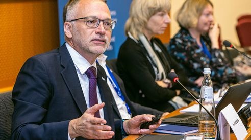A man in a suit speaks at a conference table with microphones, alongside three seated individuals, in a formal setting.