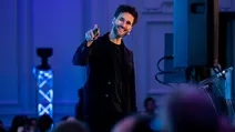 Man in a dark suit smiling and pointing while speaking on stage at an event with blue lighting.