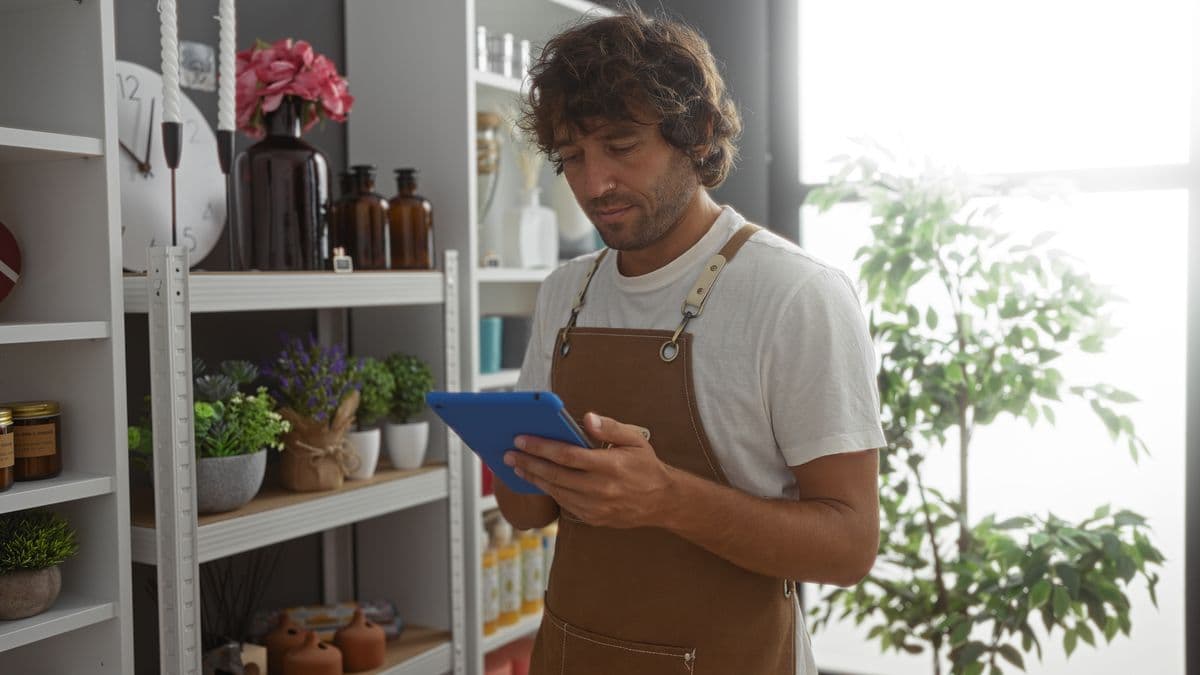 Young man in a home decor store wearing an apron, using a tablet surrounded by indoor plants and decorations