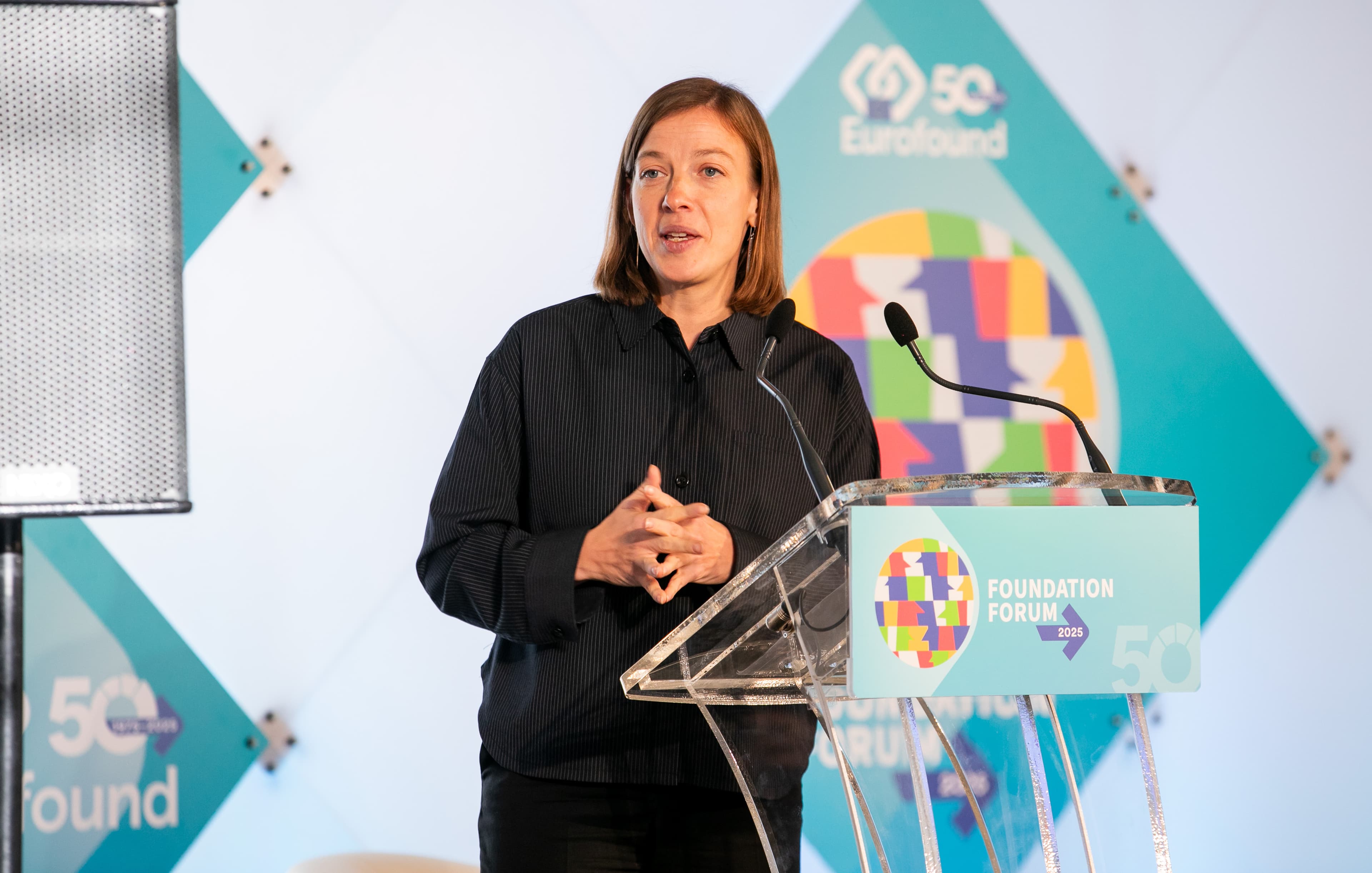 A woman speaks at a podium during the Eurofound 50th Foundation Forum, with a colorful logo and microphone in front of her.