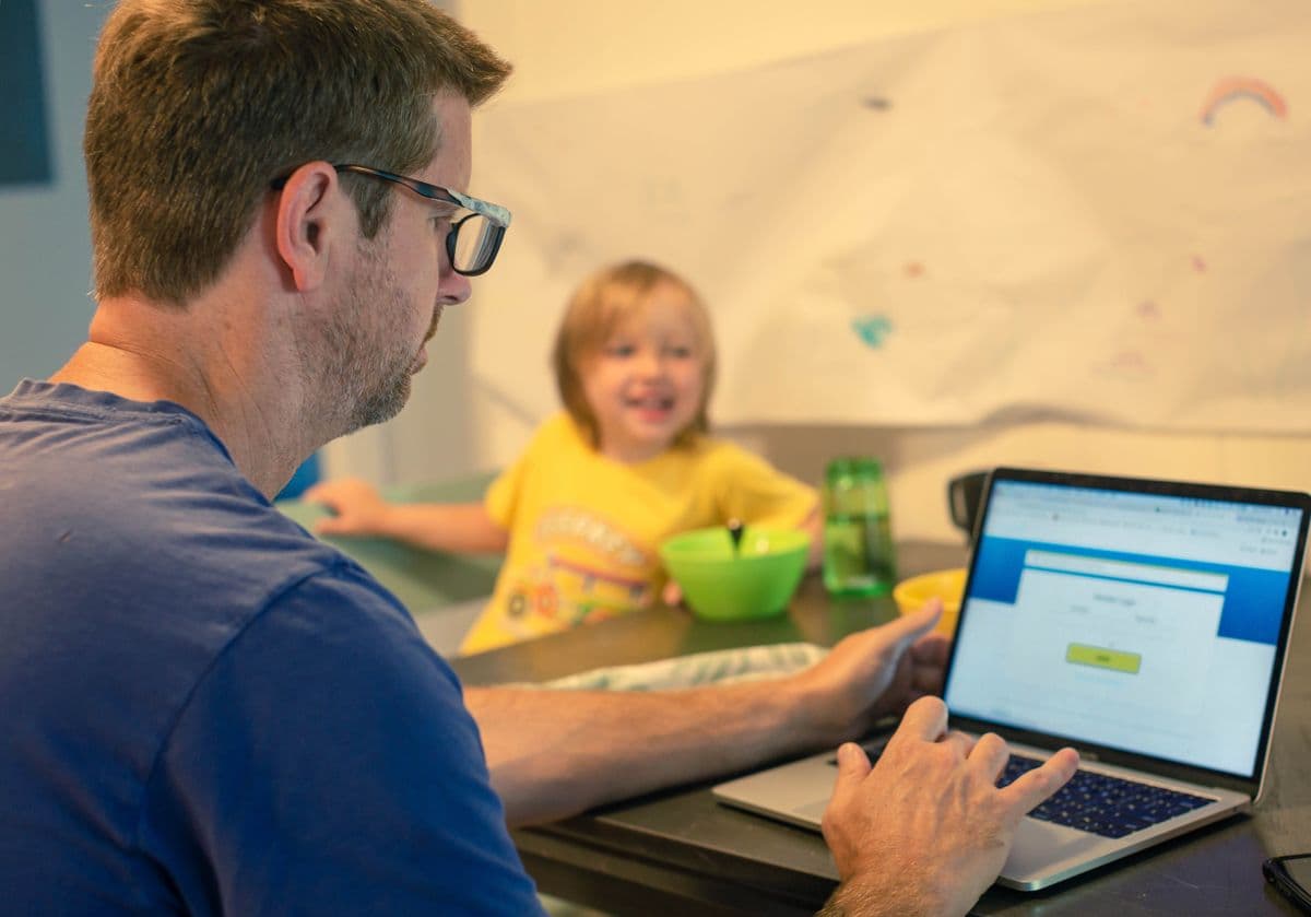 A man using a laptop at the dining table, while a child in a yellow shirt smiles in the background, sitting with a green bowl.