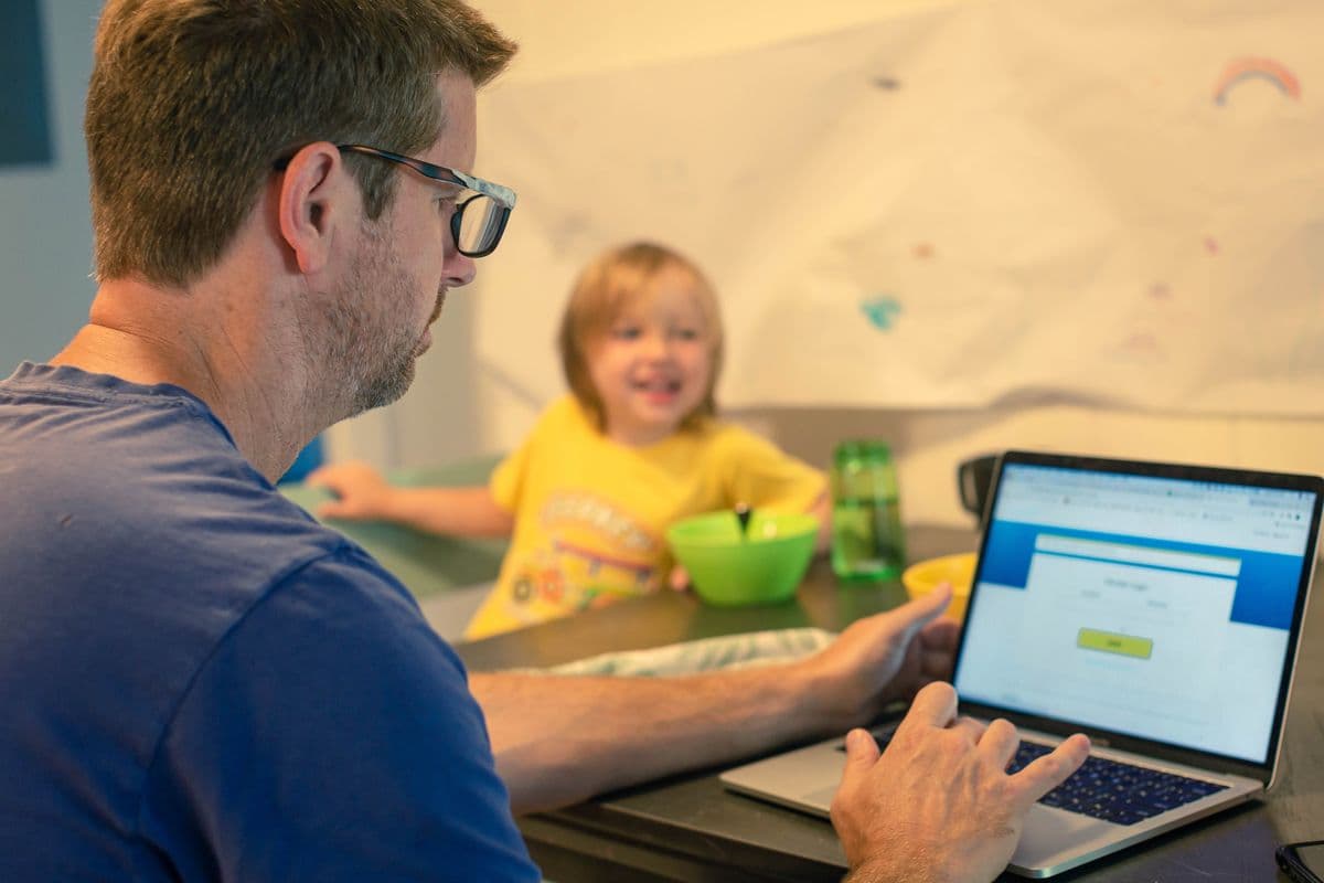A man using a laptop at the dining table, while a child in a yellow shirt smiles in the background, sitting with a green bowl.