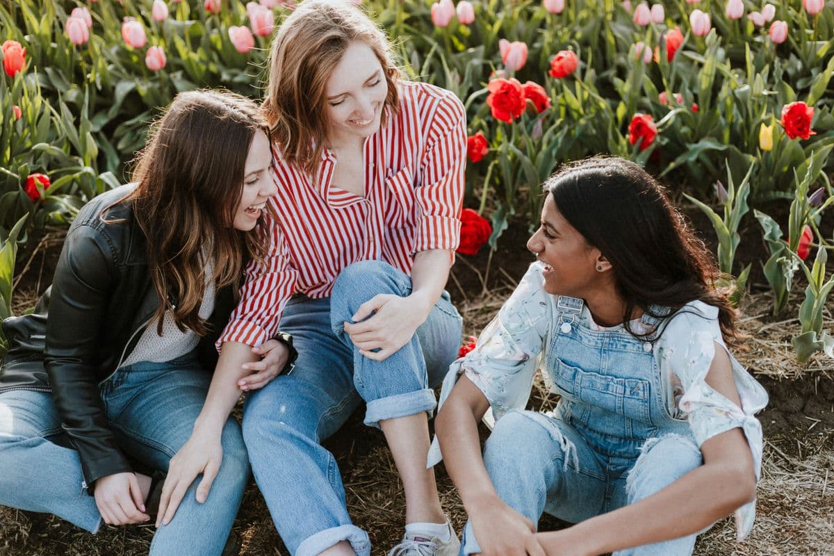 Three friends sitting and smiling together in a colorful tulip field, dressed casually in jeans and jackets, enjoying a sunny day.