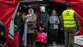 A group of Ukrainian refugees, bundled in warm clothing, stand under a red tent