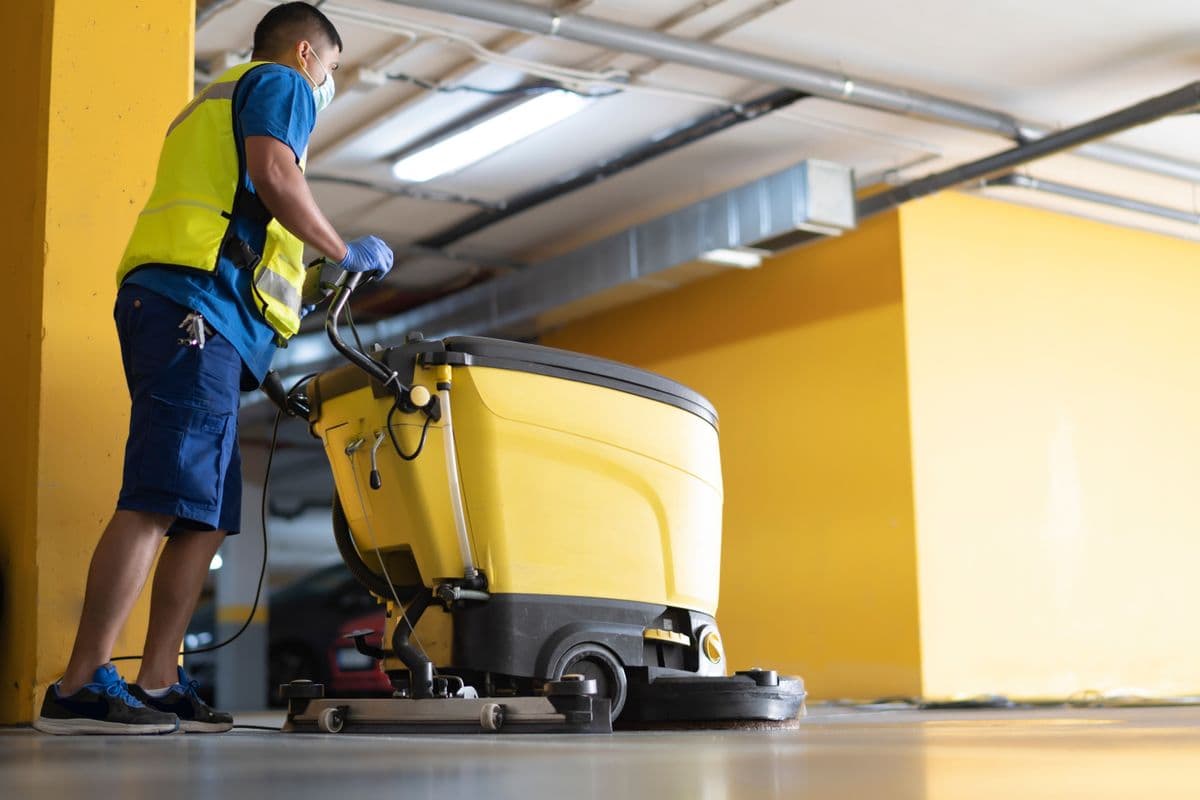 Worker in a blue uniform and safety vest operating a yellow floor cleaning machine in a parking garage.