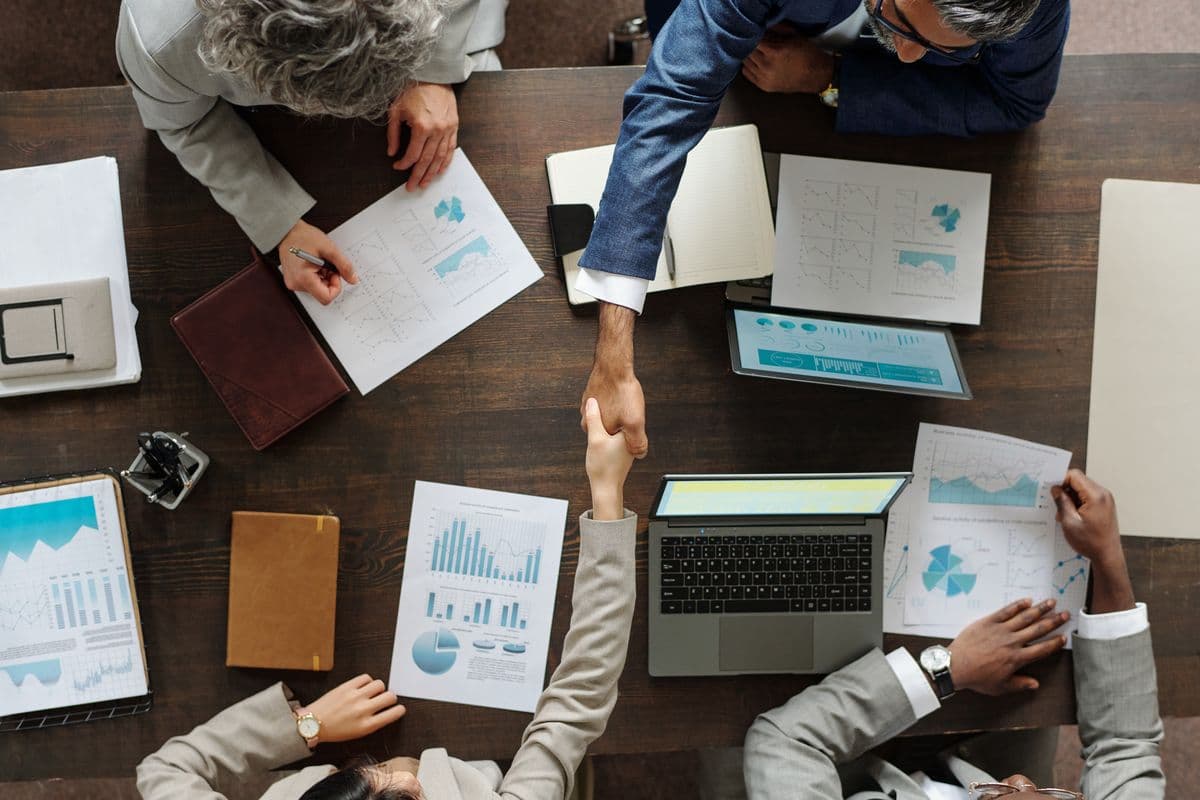Top view showing group of middle aged multiethnic business professionals collaborating at table, two people shaking hands over laptop and financial charts, others reviewing documents